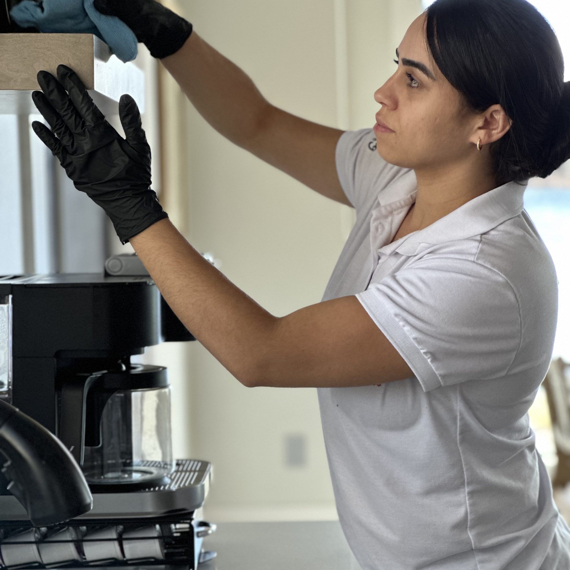A Simplify Cleaning Services professional in a pink uniform and black gloves meticulously dusting a small potted plant on a wooden shelf with a yellow microfiber cloth.