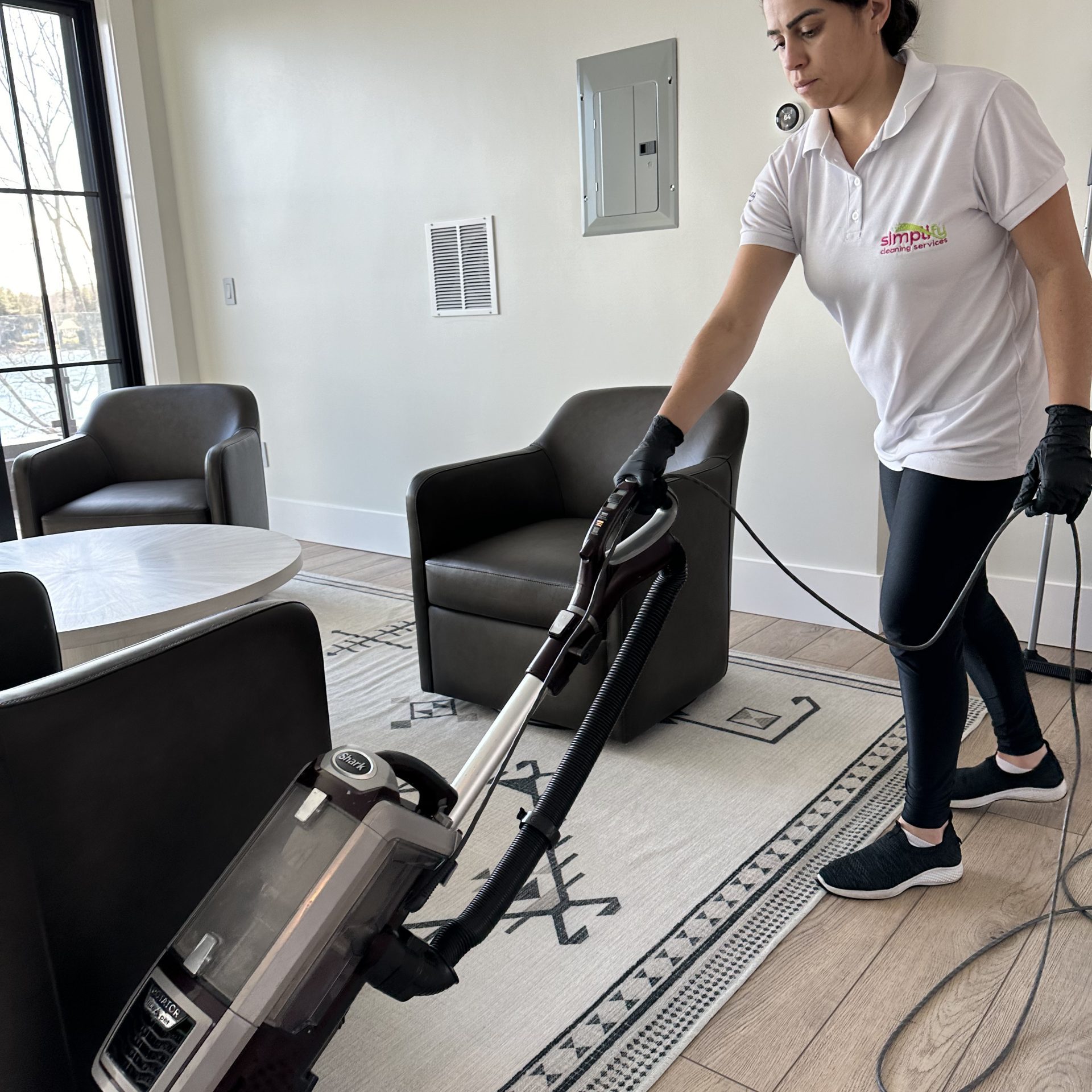 An expert from Simplify Cleaning Services using a high-quality vacuum to clean an area rug in a modern Massachusetts living room with large windows.
