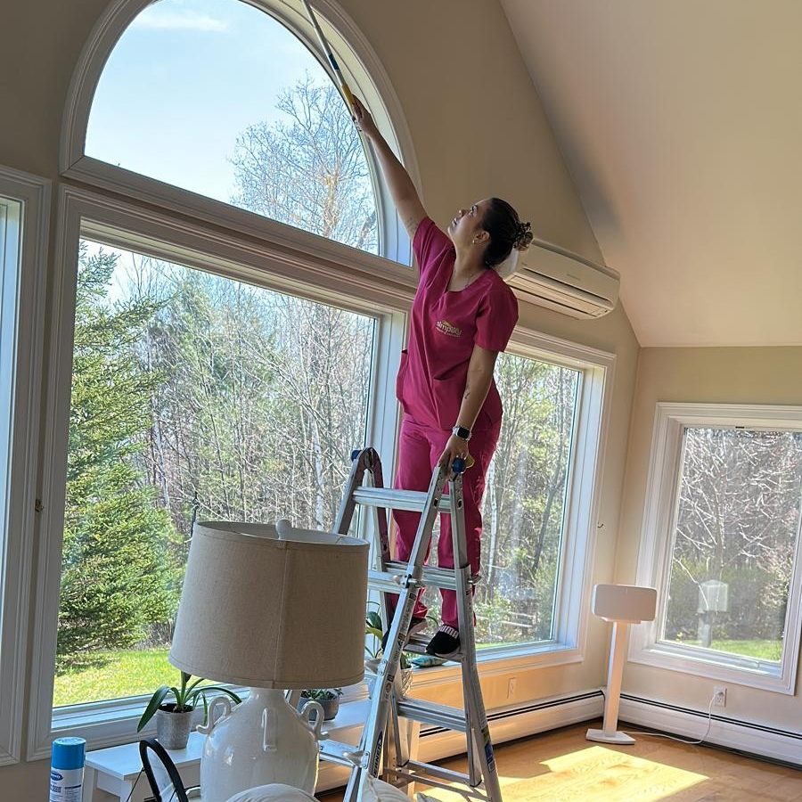 A professional cleaner from Simplify Cleaning Services in Massachusetts, wearing a branded white polo and black gloves, meticulously dusting high-reach door frames and trim.